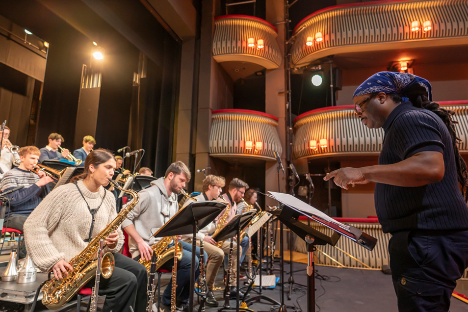 A group of students in a jazz orchestra rehearsal, performing with the conductor in a theatre.